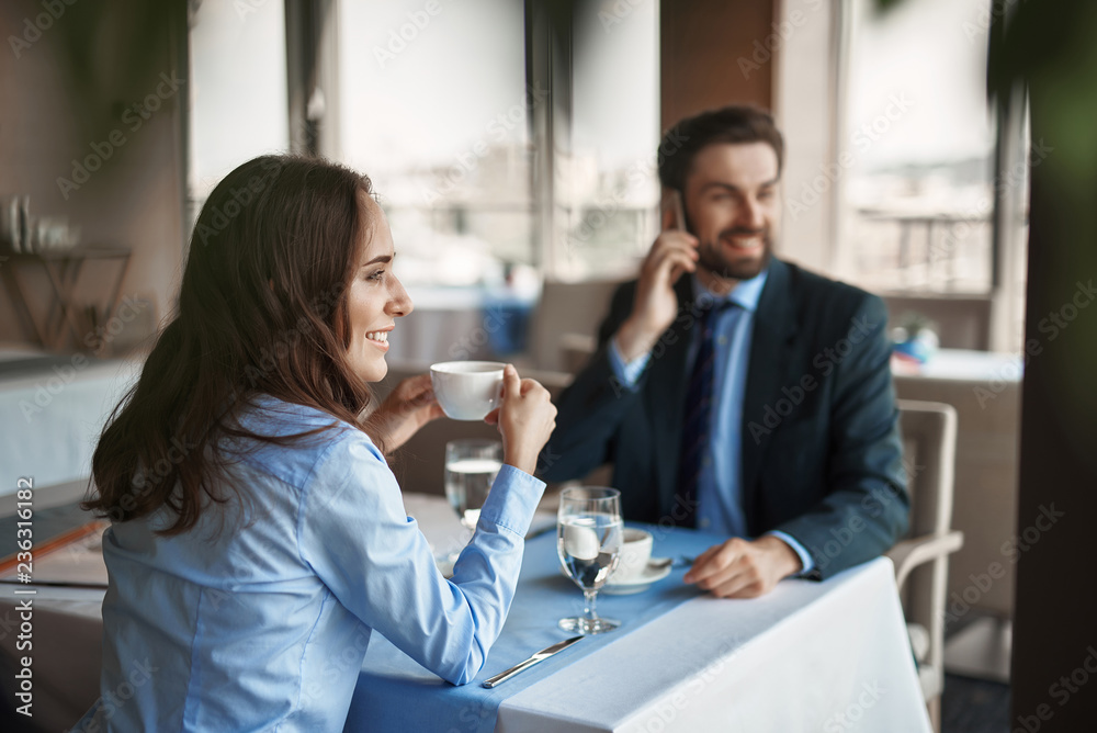 Business lunch in friendly atmosphere. Selective focus on smiling businesswoman drinking coffee while sitting in restaurant with man that talking by phone