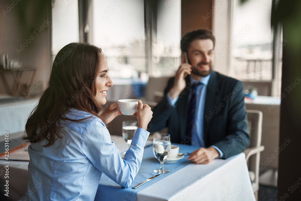Business lunch in friendly atmosphere. Selective focus on smiling businesswoman drinking coffee while sitting in restaurant with man that talking by phone Business lunch in friendly atmosphere. Selective focus on smiling businesswoman drinking coffee while sitting in restaurant with man that talking by phone