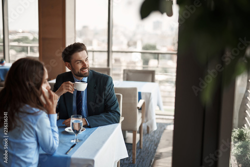 Business lunch in friendly atmosphere. Selective focus on smiling businessman drinking coffee while sitting in restaurant with woman that talking by phone