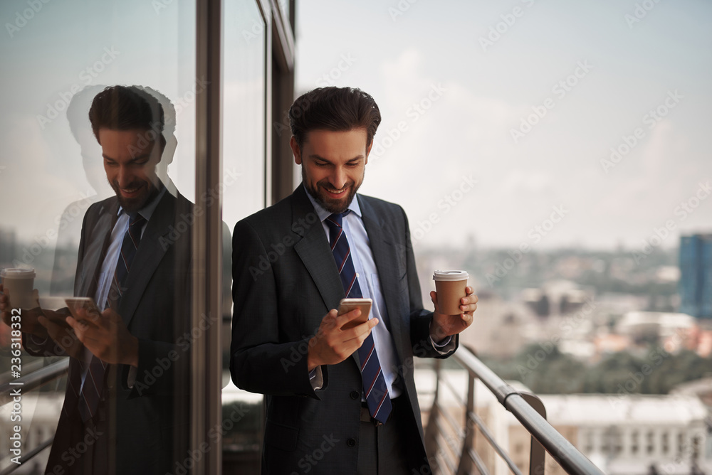 Take a pause. Waist up portrait of cheerful businessman checking massages on smartphone while standing on office balcony with cup of coffee. Copy space on right