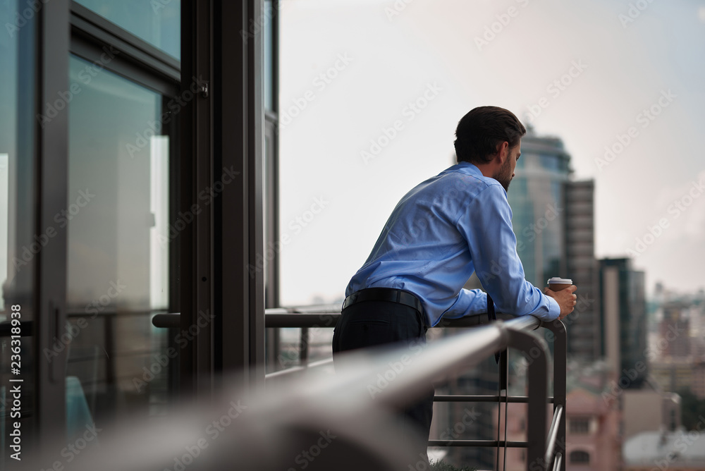 Corporate time-out culture. Back side portrait of young businessman standing on office balcony with cup of coffee and looking at city view Corporate time-out culture. Back side portrait of young businessman standing on office balcony with cup of coffee and looking at city view