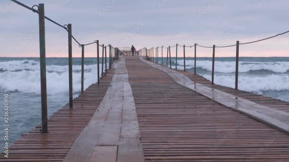 Family are walking at the sea pier in storm. Waves splashes through the wooden pier in the sea. Beautiful seascape in sunset. Family are walking at the sea pier in storm. Waves splashes through the wooden pier in the sea. Beautiful seascape in sunset.