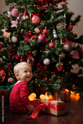 Little baby in red pajamas on the background of the Christmas tree with a gift in the festive package.