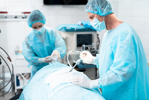 Side view portrait of middle aged doctor in protective mask and sterile gown performing surgical operation in modern operating room. Female assistant on blurred background