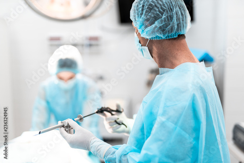 Portrait of doctor in sterile gloves holding laparoscopic grasper with trocar. He is wearing blue surgical cap and gown photo