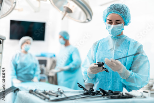 Waist up portrait of female medical worker in protective mask standing near table with surgical tools. Surgeon and his assistant on blurred background