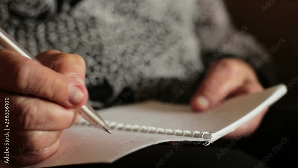 The old woman writes something in a notebook or diary. The wrinkled hand of an old woman writes something on paper