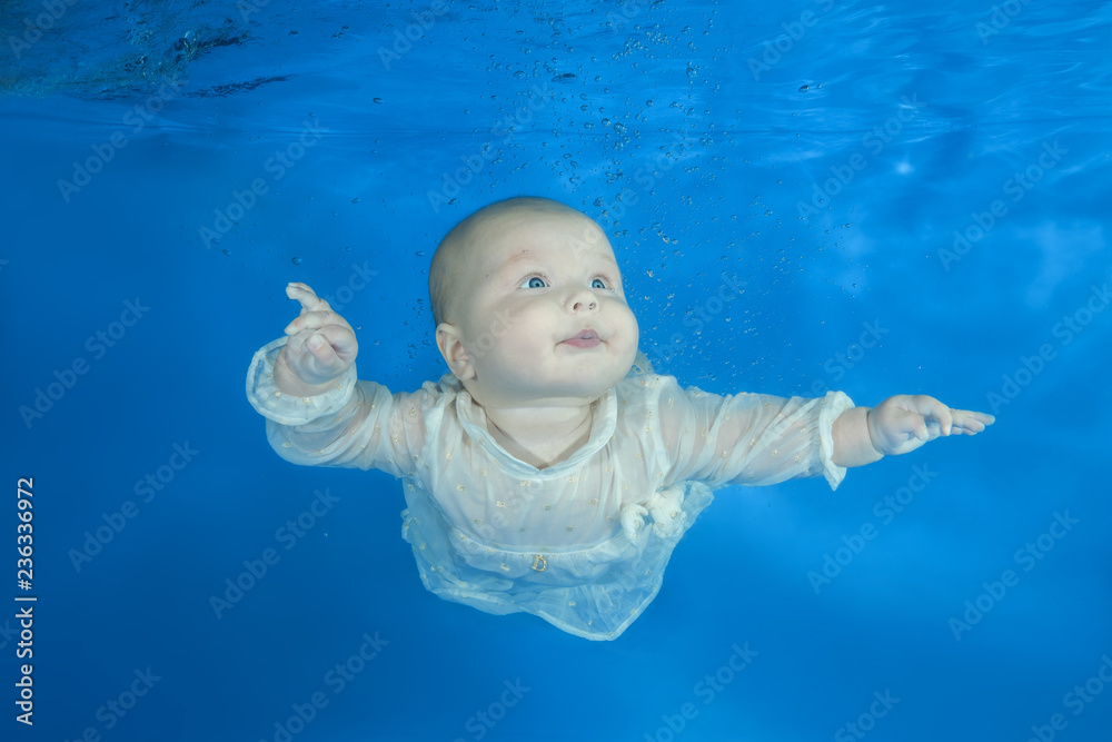 Funny face portrait of little baby girl in the white dress swim underwater on a blue water background.  Healthy family lifestyle and children water sports activity.  