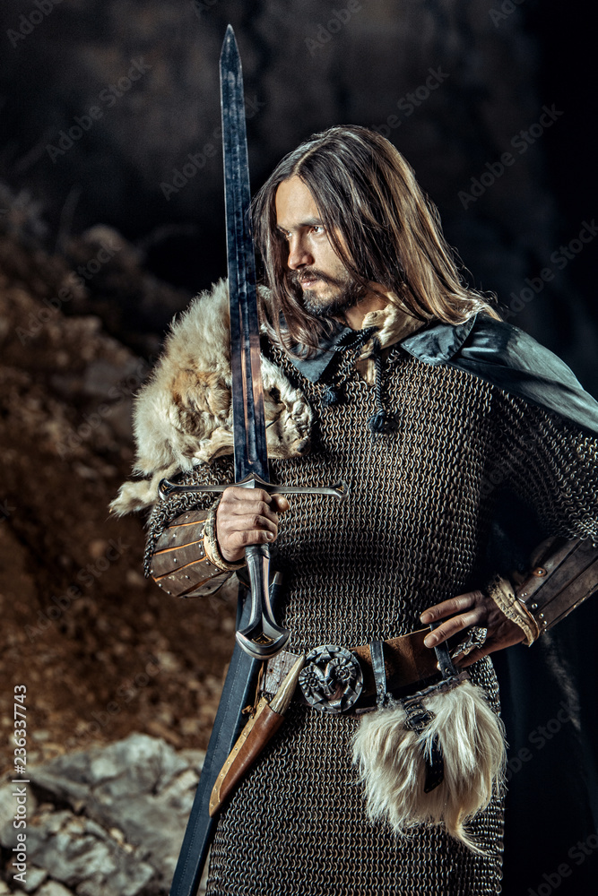 Naklejka premium Long haired knight with the two-handed sword. Rocks on the background.