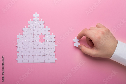 White puzzle. House shape puzzle. The concept of rent, mortgage. Hand holding piece of white puzzle. Pink background. Top view photo