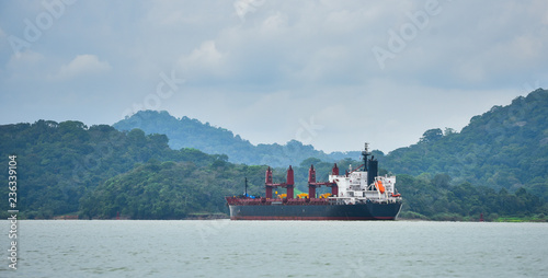 A large cargo ship makes its way through the Panama Canal waterways.  Each side between lift locks is lined with green rain forest jungle.