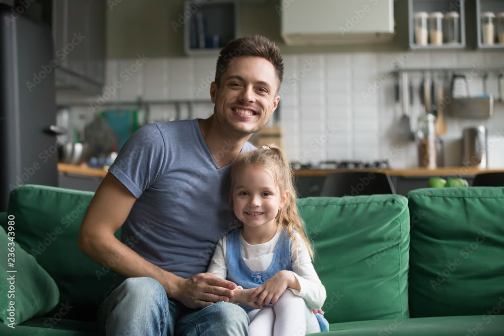 Obraz premium Portrait of happy smiling father with preschool daughter, father sitting on sofa at home together with little girl, kid sitting near dad, look at camera