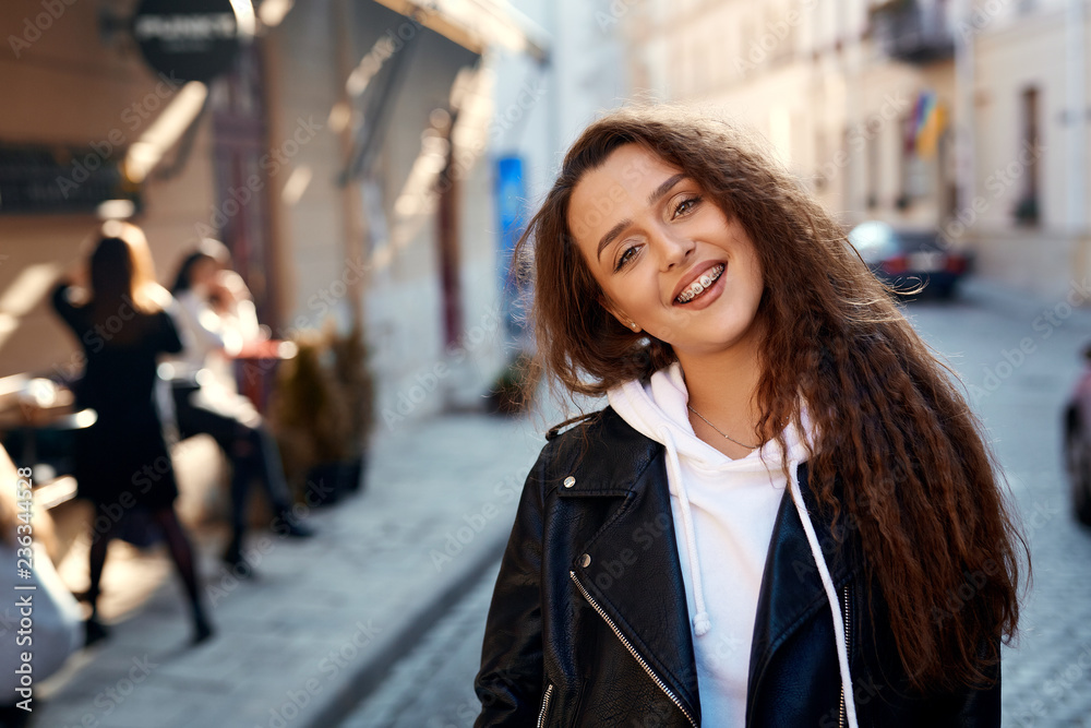 Fototapeta premium beautiful young woman walking city having fun. joyful Woman with long curly hair, wearing in black leather biker jacket and white hoodie standing on the street and smiling