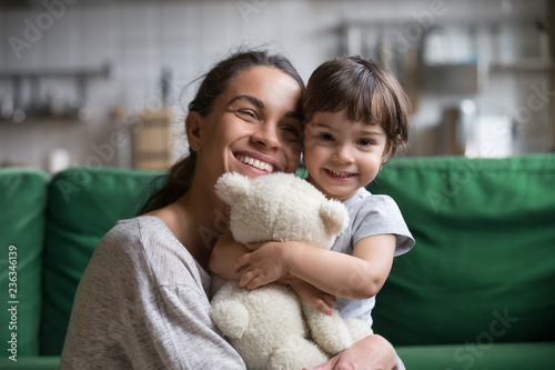 Smiling single young mum embracing little preschool daughter with toy, playing in living room at home, mother laughing with child, headshot portrait, cute girl look at camera