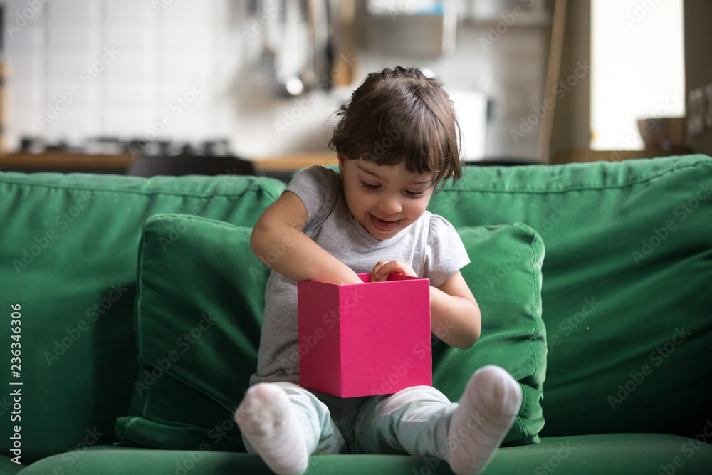 Excited curious little preschool cute girl opening, checking gift box, sitting on sofa, couch, receiving presents, looking inside opened pink box, celebrating birthday at home, got new toy Excited curious little preschool cute girl opening, checking gift box, sitting on sofa, couch, receiving presents, looking inside opened pink box, celebrating birthday at home, got new toy
