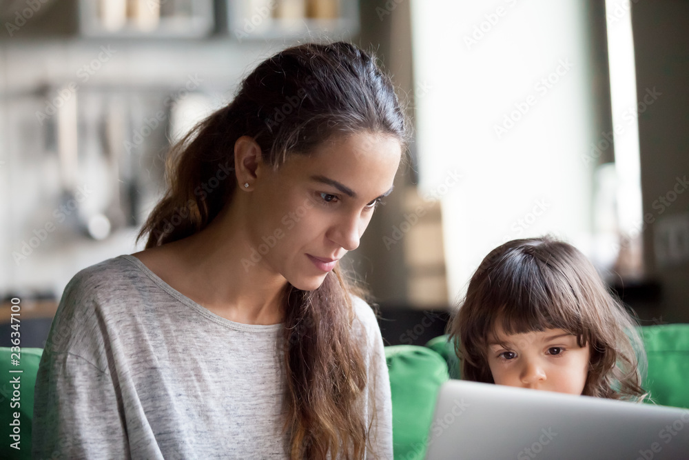 Serious focused single mother using laptop together with little preschool daughter, girl watching working mum, babysitter searching cartoons for pupil, showing training materials Serious focused single mother using laptop together with little preschool daughter, girl watching working mum, babysitter searching cartoons for pupil, showing training materials