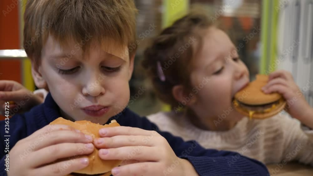 Hungry children greedily eating burgers boy and girl sit in fast food restaurant Hungry children greedily eating burgers boy and girl sit in fast food restaurant