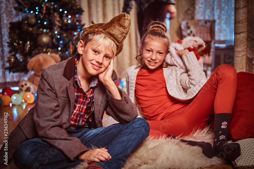 Happy brother and sister sitting on a fur carpet near a Christmas tree at home.