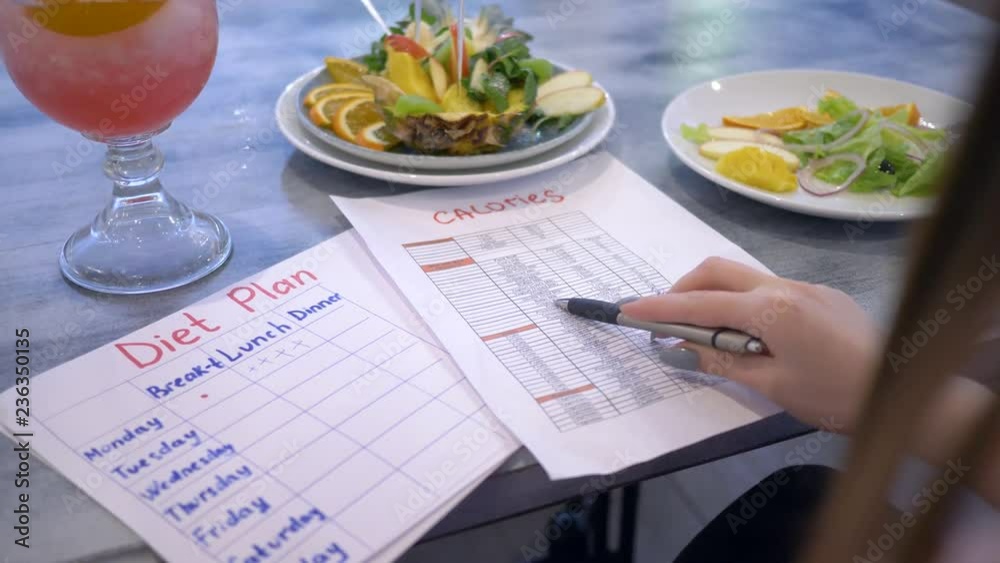 nutritionist female looking for calories in the list and hold in hand diet plan sitting at the table with punch and fruits in plate