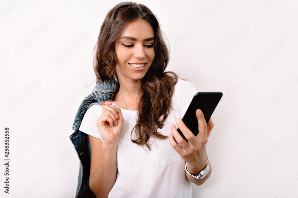 Close up portrait of cheerful beautiful girl with dark long hair in casual white shirt smiling with teeth, looking at her smartphone with happy and relaxed face expression Close up portrait of cheerful beautiful girl with dark long hair in casual white shirt smiling with teeth, looking at her smartphone with happy and relaxed face expression