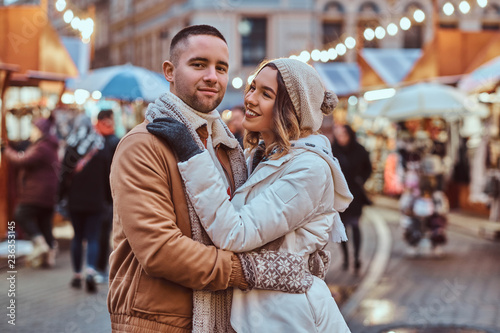 A young romantic couple wearing warm clothes hugging outdoor in evening street at Christmas time, enjoying spending time together.