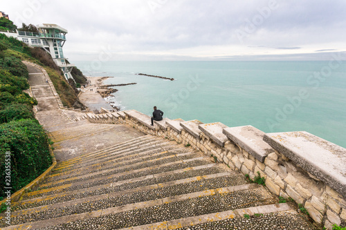 Fototapeta Naklejka Na Ścianę i Meble -  Stairway Passetto Ancona Marche Italy with man sitting on the stairway thinking and watching the sea. The Passetto is a panoramic point over the Adriatic Sea, with concrete stairway symbol of the city