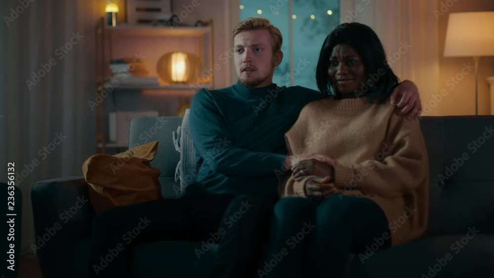 Happy Young Couple Watching Horror Movie on TV while Sitting on a Couch, they got Scared, Girl Covers eyes. Handsome Caucasian Boy and Black Girl in Love Spending Time Together. Moving Camera Shot.