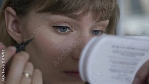 Young beautiful blonde woman applying eyeliner doing perfect eye arrows professional trendy makeup indoors.