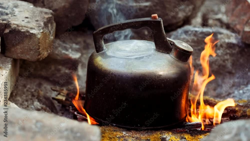 Dolly shot of old metal kettle on camping fire with boiling water. Traveler making tea with teapot on picnic in forest, smoke comes from campfire. Hiker with travel pot outdoors on nature. Dolly shot of old metal kettle on camping fire with boiling water. Traveler making tea with teapot on picnic in forest, smoke comes from campfire. Hiker with travel pot outdoors on nature.