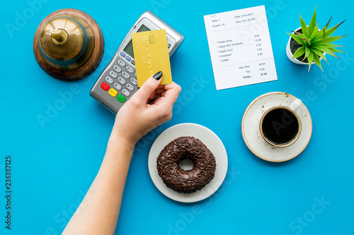 Pay the bill by payment terminal. Woman's hand insert bank card in payment terminal near bill, service bell, coffee and donuts on blue background top view copy space