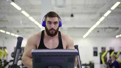 professional athlete bodybuilder performs cardio training on the ellipsoid simulator in the gym. listens to music through wireless headphones and drinks milk protein