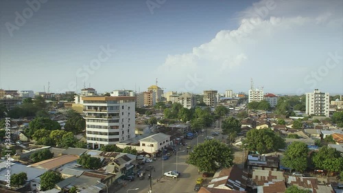 Wide still shot,  aerial view, calm morning,  Conakry city highway road, clean,  streetlights, low traffic, people walking, leafy trees,  rusty roofs,  few modern flats, white blue clouds, ho photo