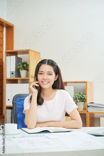 Portrait of smiling lovely female entrepreneur sitting at office table and filling her planner
