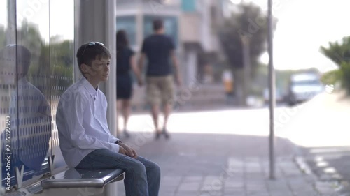 boy sits on the bus stop and waits for the bus, looks at watch