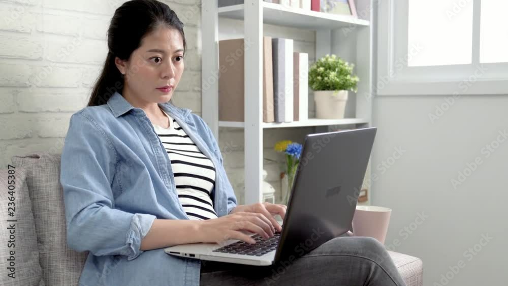 beautiful woman on laptop computer typing project drinking tea in the morning before going to work. young asian lady working at home office on comfortable couch in bright living room sunlight window.