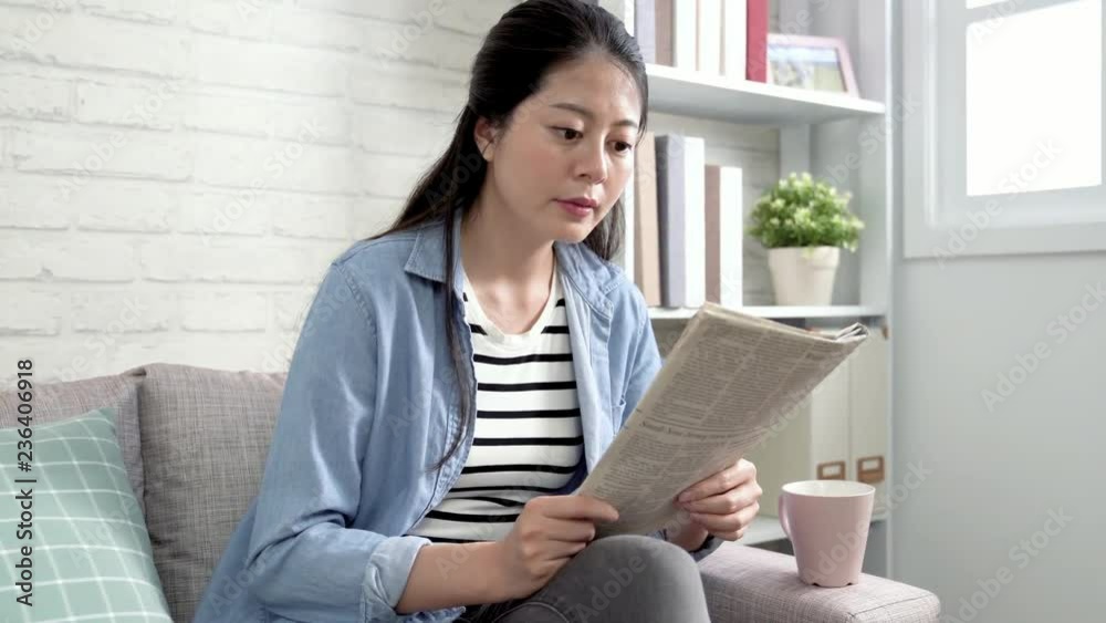 slow motion young asian wife relaxing in sofa concentrated reading newspaper drinking hot coffee in afternoon tea time in living room in apartment. beautiful mother interesting about daily article.