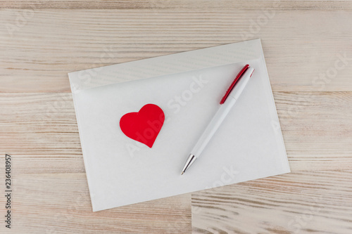 любовная почта: белых пустых незаполненных конверт для писем, белая шариковая ручка и красное сердечко лежит сверху, светлый деревянный стол