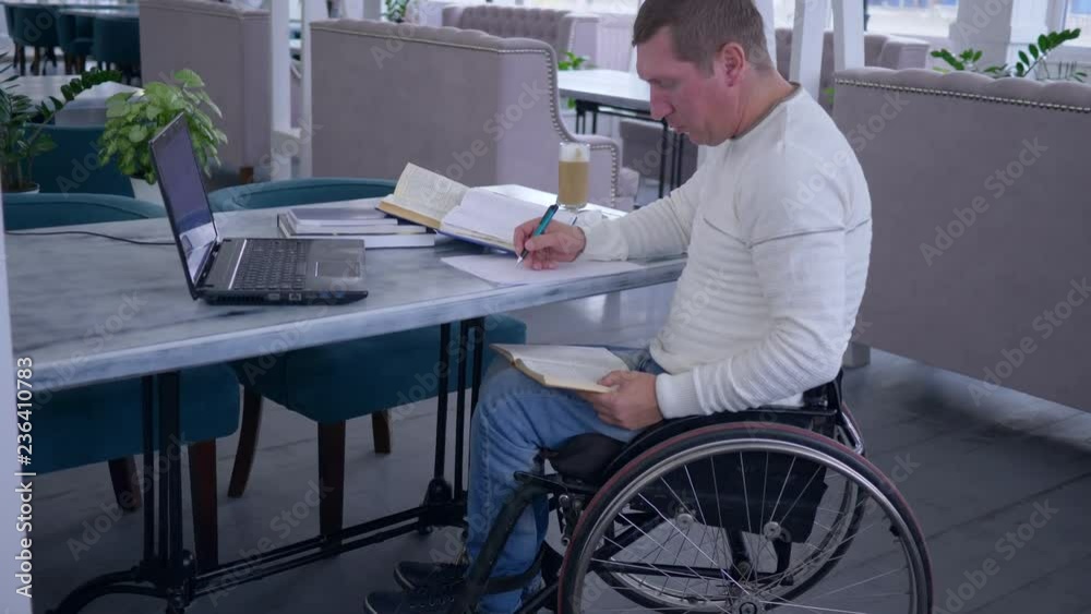 successful diseased student man on wheel chair works on laptop computer during distance learning for online education and makes notes in notebook in restaurant