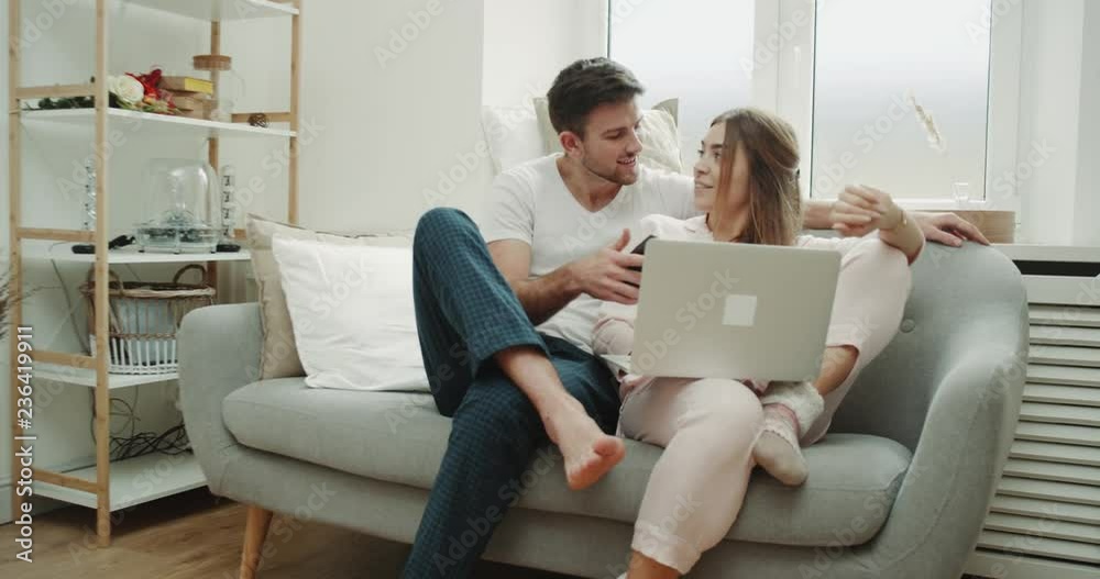 In the morning couple in pajamas in a stylish living room ordering something from the phone , and using notebook by the way.