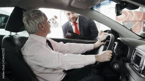 Sales man is giving an overview of the car. Business man is in the car, listening and trying the steering wheel.