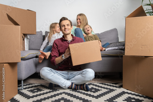 Image of happy man sitting on floor and women with boy and girl sitting on gray sofa among cardboard boxes