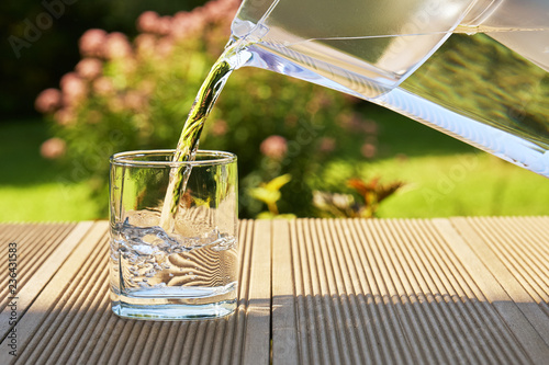 Pouring clear filtered water from a water filtration jug into a glass in green summer garden in a sunny summer day photo