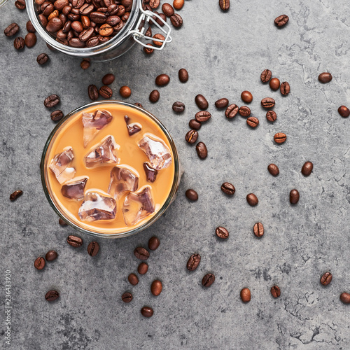 Chocolate, vanilla, caramel or cinnamon iced coffee in tall glass. Cold brewed iced coffee in glass and coffee beans in glass jar on grey concrete background. Top view, square crop.