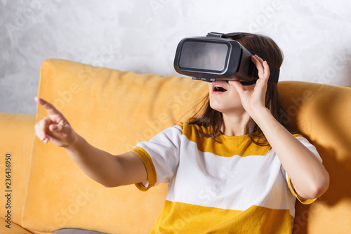 Surprised long-haired girl in pullover touching something with her hands while testing VR devicee, she sitting on the yellow sofa in the light living room, future technology concept