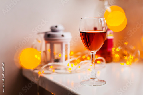 A glass of red wine on the white table with decor lantern and blurred bokeh lights on the background. Concept of cozy atmosphere and ambience at home. Warm colors
