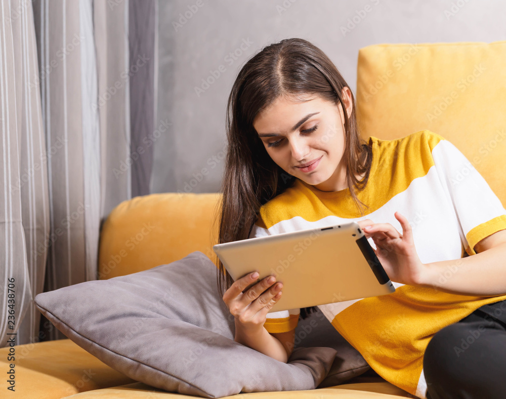 Cute long haired brunette girl laying on the yellow sofa in the light living room and browsing Internet on the digital tablet, modern technology, communication concept Cute long haired brunette girl laying on the yellow sofa in the light living room and browsing Internet on the digital tablet, modern technology, communication concept