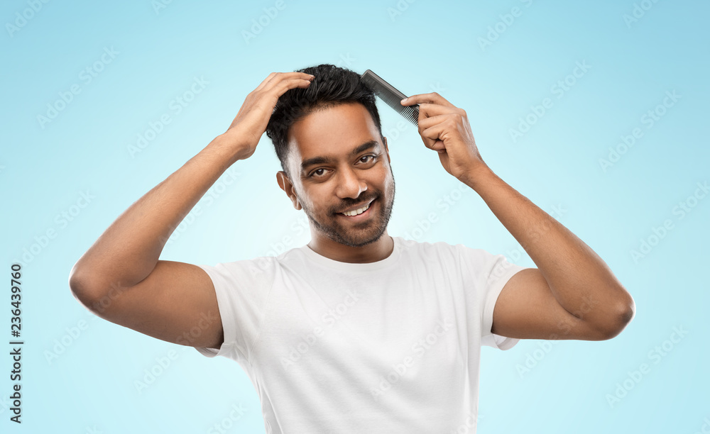 Fototapeta premium grooming, hairstyling and people concept - smiling young indian man brushing hair with comb over blue background