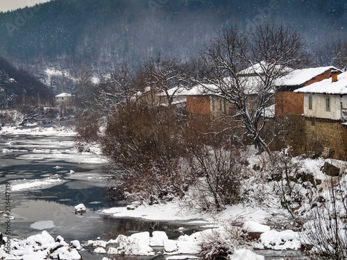 Picturesque elevated snowy rural view from a bridge to the winter Vidima River and old riverbank houses at the Bulgarian village of Debnevo, Central-North Bulgaria photo