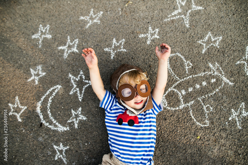 Portrait of smiling toddler wearing pilot hat and goggles lying on asphalt painted with airplane, moon and stars photo