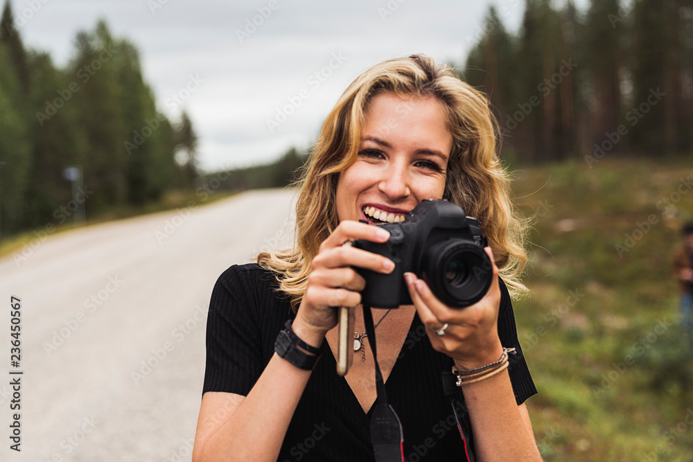 Finland, Lapland, portrait of happy young woman holding camera at country road Finland, Lapland, portrait of happy young woman holding camera at country road